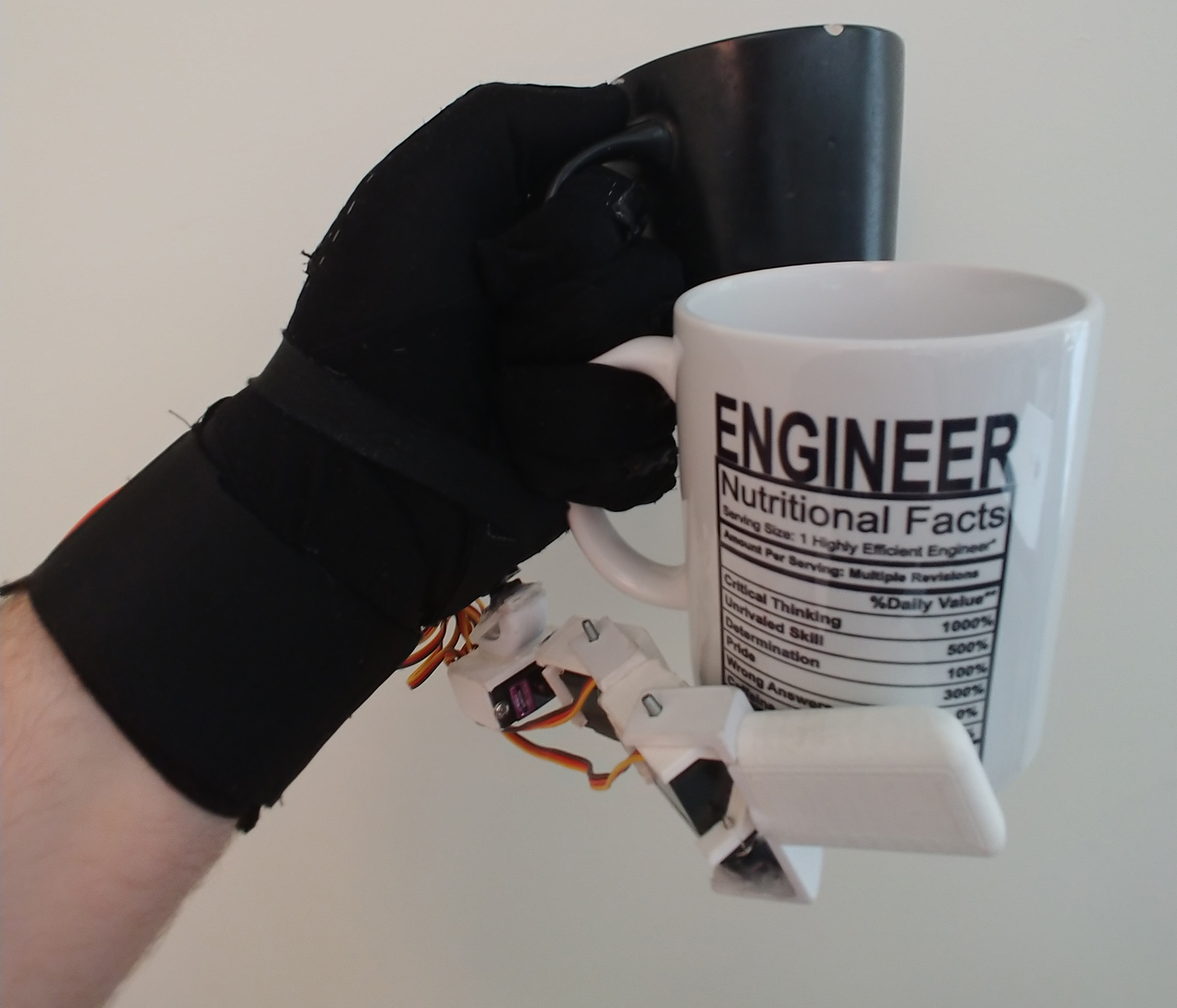 Picture shows a gloved hand holding a cup with thumb, index finger, and middle finger. A second cup is being held with the ring finger, pinky, and the supernumerary robotic opposable finger